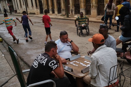 Four men play dominoes on a table in the street. Other people including children and a man in a wheelchair can be seen in the street behind them