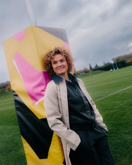 Ellie Kildunne with her hands in her pockets leaning against pink, yellow and black padding round a rugby post, wearing a white vest underneath a black tracksuit with a beige trenchcoat over the top