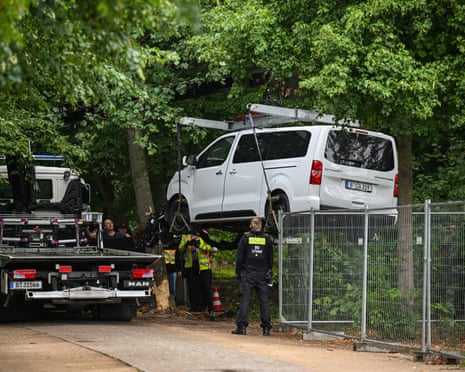 A white Citroen passenger van is lifted over wire fencing towards a tow truck on a tree-lined path as police stand by.