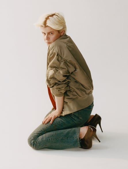 روبن مكتوفة على ركبتيها مع جسمها متجهًا نحو اليمين، يديها على فخذيها، ترتدي سترة بومبر ذات لمعة خفيفة مع الأكمام المطوية، جينز قديم وحذاء كعب عالٍ برونزي، تنظر إلى الكاميرا