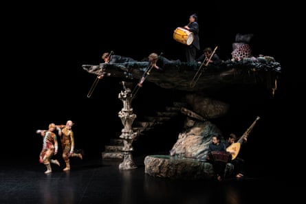 Two dancers on stage beside set of bony granite with musicians on top of and below it
