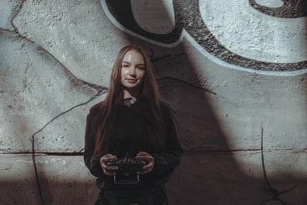 A young woman with long hair stands in front of a graffitied wakk holding a control panel