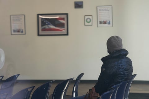 a person sits in a row of empty plastic chairs in a room where a picture of the US flag is hung on the wall