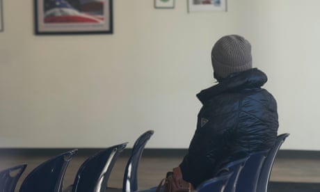 a person sits in a row of empty plastic chairs in a room where a picture of the US flag is hung on the wall
