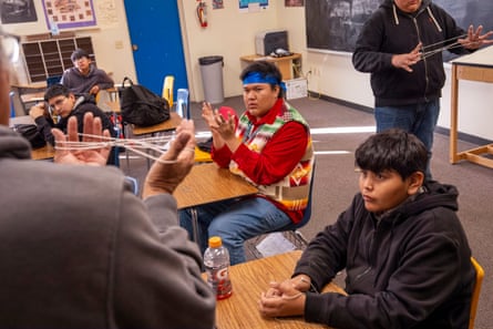 Students sitting at desks watching someone play a game like cat’s cradle with string in their hands