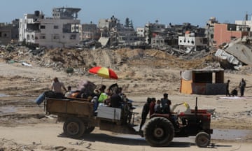 Palestinins ride with their belongings on a trailer pulled by a tractor, with destroyed and damaged buildings in the background