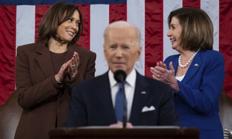 Kamala Harris and Nancy Pelosi smiling at each other behind Joe Biden