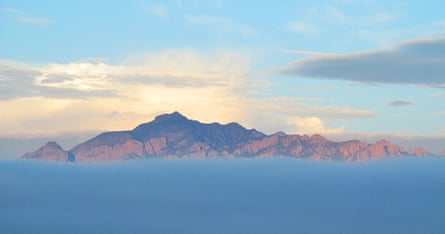 Mountain in distance rises above thick layer of clouds under blue cloudy sky