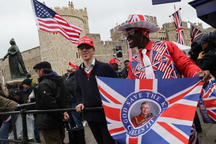 A black man dressed in a red suit with lapels decorated with the US and UK flags and a hat and sunglasses decorated with the union jack, holding a flag declaring ‘God Save the King Charles III’ stands beside a white in a dark navy fleece wearing a red Make America Great Again cap