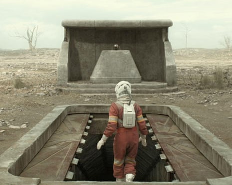 A person in a space suit steps into a bunker surrounded by a wasteland.