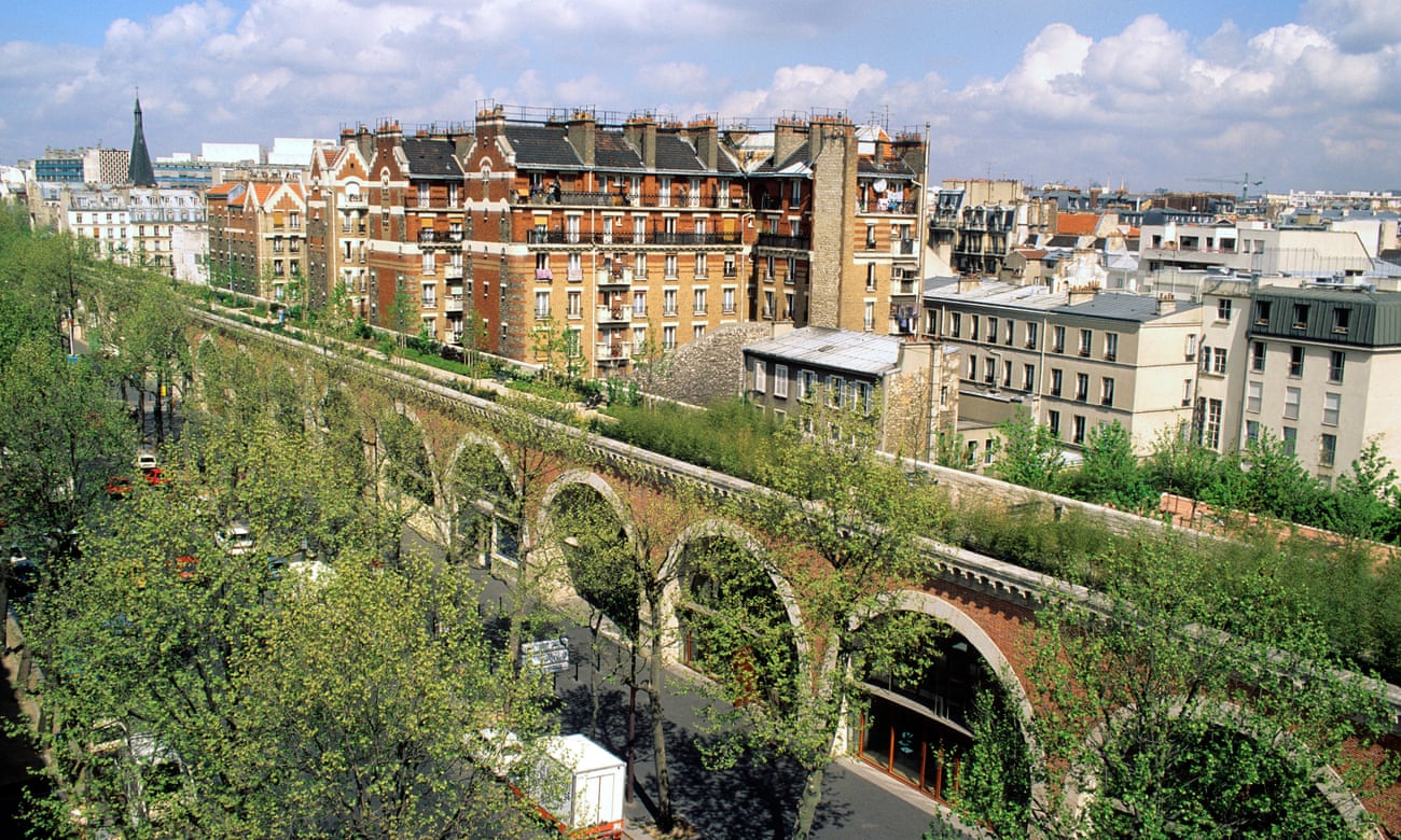 The image shows the  Promenade Plantée, an park built on an abandoned viaduct. The stricture resembles an elevated train track, made of brick arches, with shop fronts in the space under the arches. The top of the viaduct has grasses, shrubs, and benches, with trees on either side of it, and the buildings of Paris in the background