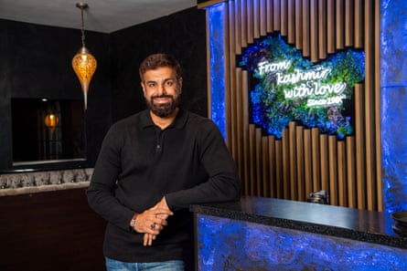 He leans against a wall in his restaurant, which is lit in blue light. A sign reads ‘From Kashmir with love’