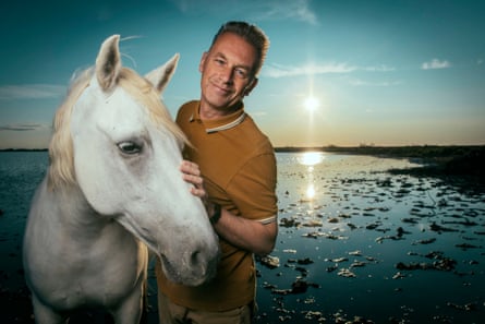 Chris Packham strokes a white horse with a sunset over the sea in the background