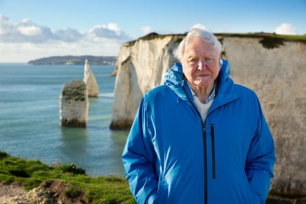 An elderly man in a blue waterproof jackets stands on a hilltop with the sea and the white cliffs of Dover behind him.