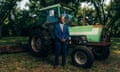 A man wearing a blue suit stands in front of a green tractor in a grassy field.
