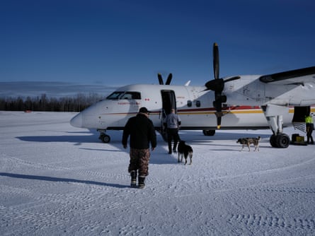 John Meeseetawageesic walks across a snow-covered runway to board a small prop plane. A man and two loose dogs walk ahead of him. He wears snow boots and heavy trousers.