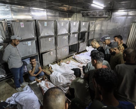Mourners survey a row of bodies wrapped in sheets lain on the floor of a mortuary room