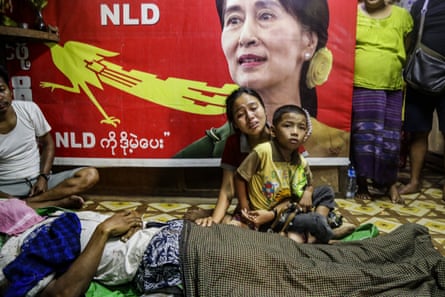 A woman weeps while holding a child next to the covered body of a protester. In the background is a banner of Aung San Suu Kyi.