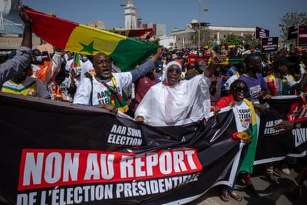 Protesters waving flags and placards march in the street behind a banner saying; ‘Non au report de l’élection presidentielle’
