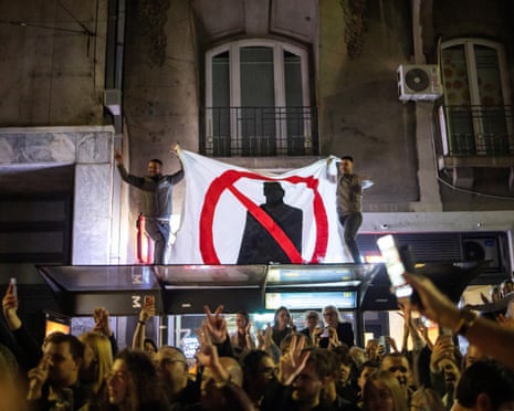 Crowd in Belgrade gathers at night as men display an anti-Vučić banner from a rooftop