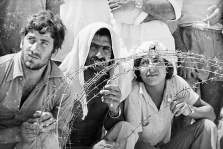 A bearded man in a collared shirt with buttons undone sits next to a bearded man with a cloth over his head and a young man in a short-sleeved shirt, all squinting into the sun behind strands of barbed wire on Cyprus in 1974