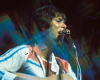 Cliff Richard sings into a microphone while playing guitar on stage