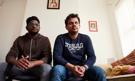 Akhil Jenny and Geo Ambooken: they are sitting on a couch in a white-painted and rather bare-looking living room with a light brown sofa and striped cushions. Jenny wears a brown zip-up hoodie and Ambooken, who also has short black hair and a beard, wears a dark blue sweatshirt with a logo in white reading Staywild and blue jeans.