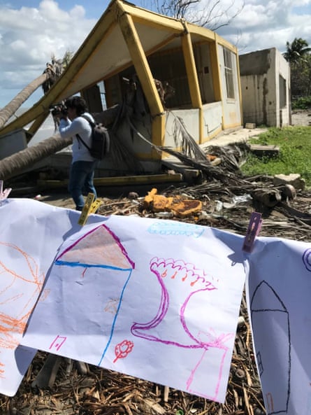 Children’s drawings of houses hang on a line, attached with pegs, with fallen trees and damaged buildings in the background