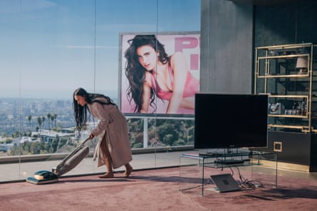 Woman vacuuming in a robe with a billboard behind a glass wall overlooking the city
