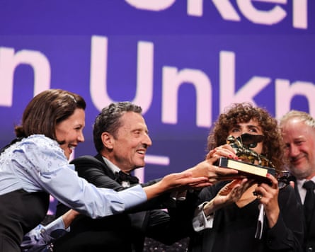 Maggie Gyllenhaal laughing and Pietrangelo Buttafuoco smiling as they both lean towards May el-Toukhy with the Golden Lion