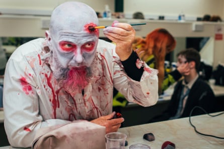 A man in white face paint and a boilersuit paints blood on to his forehead.
