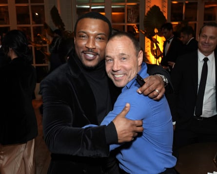 The two men smile, looking into the camera, with their arms around each other