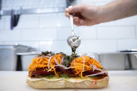 A chef’s hand spooning salsa verde on to a salad sandwich in the making.