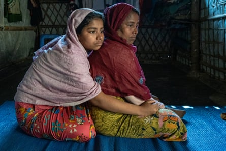 Two women sit on a mat, the younger hugging the elder from behind.
