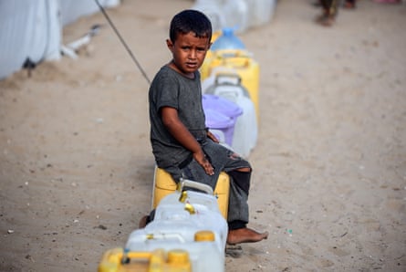 A child sits on a row of water butts lined up on the sand.