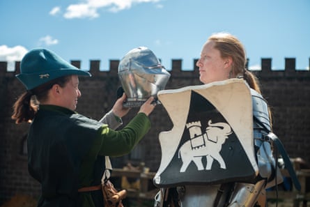 A young woman in a green medieval outfit and cap holds up a helmet to put it on a young woman wearing a suit of armour and holding a shield.