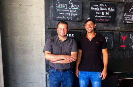 Two men in jeans and polo shirts stand in front of a chalk noticeboard