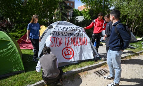 five young men are erecting a tent on the grass outside a campus building, and have draped a banner over it which reads Stop Genocide. They are dressed in T-shirts, sweatshirts and jeans and the sun is shining; other tents are close by on the grass.  eiddirdiqteinv