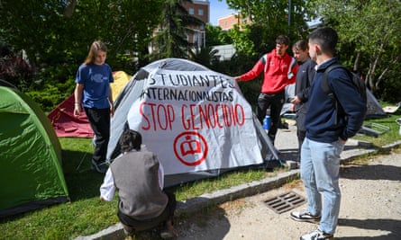 five young men are erecting a tent on the grass outside a campus building, and have draped a banner over it which reads Stop Genocide. They are dressed in T-shirts, sweatshirts and jeans and the sun is shining; other tents are close by on the grass.