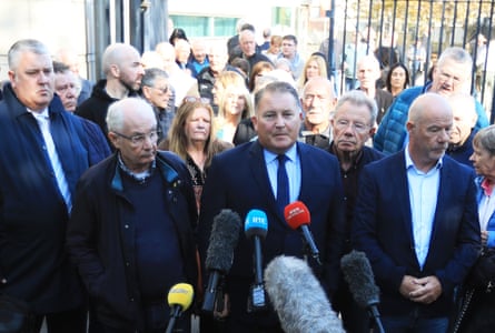 he wears a dark blue suit and is facing microphones; he stands amid a crowd of men and women of all ages, sombrely dressed.