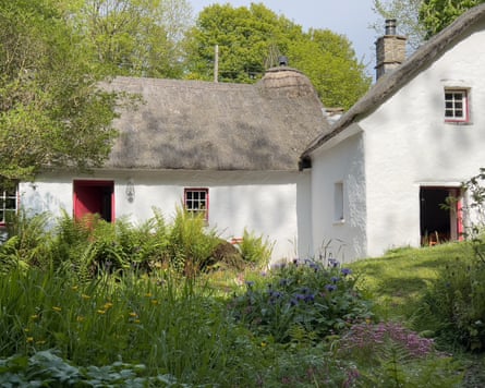 Glan Yr Afon, a white thatched cottage with open red doors, with a cottage garden in front and trees behind.