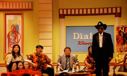 A group of people sitting in a bright yellow room. One man is wearing a black suit, white shirt and a cowboy hat with a sheriff star