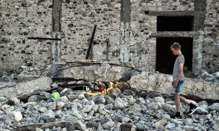 A boy stands next to a car crushed under rubble on Kos