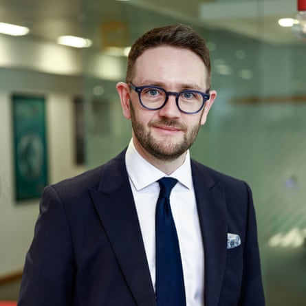 Chris Stark: portrait in an office. He wears a dark suit and tie with a white shirt and dark-rimmed glasses, and has short, light brown hair