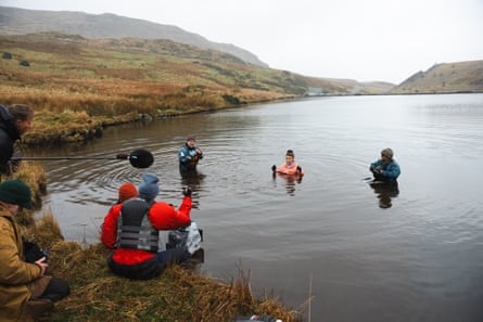 A film crew, some in the water and some on the verge, direct a woman in a lake. She is up to her chest in the water