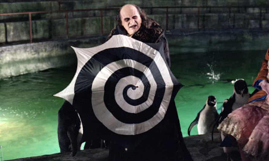 Danny DeVito plays The Penguin, a character with a pale face and beak-like nose. In this scene, he is standing in streams surrounded by penguins and holding an open swirl-shaped black and white canopy as a shield.