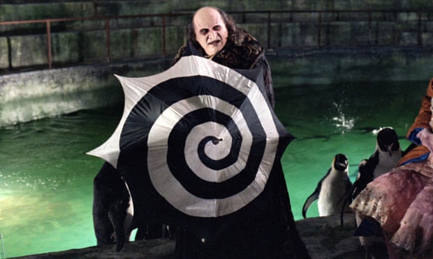 Danny DeVito plays The Penguin, a character with a pale countenance and beaklike nose. In this scene, he stands in a sewer surrounded by penguins and holding an open umbrella with a black and white swirl pattern as a shield.