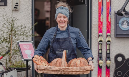 A woman, grinning, holding a flat basket of loaves of bread outside KJ’s Bothy Bakery