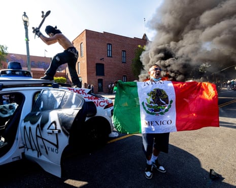 A demonstrator hits a Waymo self-driving car with his skateboard as another burns in the background, during a protest against federal immigration sweeps