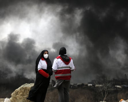 Two women in veils and firefighting uniform stand in front of a sky filled with black smoke